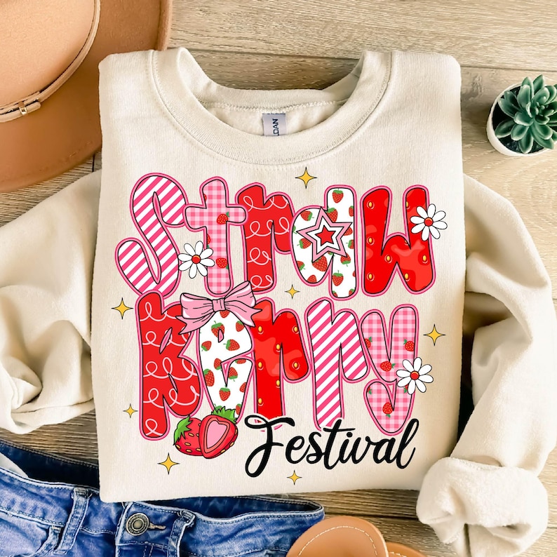 May include: A cream-colored sweatshirt with a pink and red graphic that says "Strawberry Festival". The graphic features strawberries, stars, and flowers.