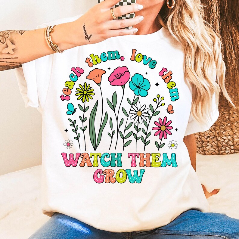 Puede incluir: Camiseta blanca con un dise&ntilde;o gr&aacute;fico colorido con flores y el texto "teach them, love them, watch them grow".