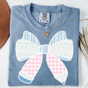 May include: A blue t-shirt featuring a large patchwork bow design in pink, blue and white. The bow is made from different fabrics, including gingham, floral and striped patterns. The shirt has the text "COMFORT COLORS" on the tag.