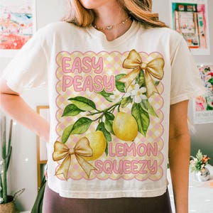 Pu&ograve; includere: Una maglietta bianca con un design grafico rosa e giallo. Il grafico presenta limoni, foglie e fiocchi. Il testo sul grafico dice "Easy Peasy Lemon Squeezy".