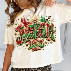 Pode incluir: Camiseta creme com um design colorido e com joias "Juneteenth 1865". O design inclui detalhes em vermelho, verde e preto, juntamente com uma paisagem urbana e estrelas. A camisa &eacute; um estilo casual de manga curta.