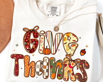 May include: Off-white t-shirt featuring the phrase "GIVE THANKS" in a colourful autumn design. The letters are decorated with leaves, pumpkins, and seasonal patterns. A gold necklace with a pendant is draped across the shirt.