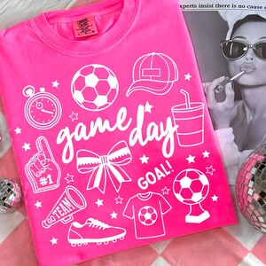 Może przedstawiać: Jasnor&oacute;żowa koszulka z białymi elementami graficznymi, w tym piłką nożną, czapką, stoperem, kokardą oraz napisami "game day" i "goal". Koszulka zawiera r&oacute;wnież koszulkę piłkarską i megafon.