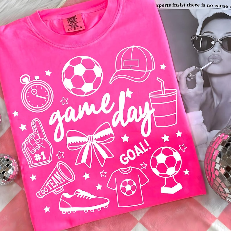 Może przedstawiać: Jasnor&oacute;żowa koszulka z białymi elementami graficznymi, w tym piłką nożną, czapką, stoperem, kokardą oraz napisami "game day" i "goal". Koszulka zawiera r&oacute;wnież koszulkę piłkarską i megafon.