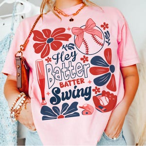 May include: Pink t-shirt with a baseball theme. The shirt features a baseball, bats, flowers, and the text "Hey Batter Batter Swing".