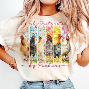May include: Cream-colored t-shirt featuring a colorful floral design with six chickens. The text "Totally Distracted by Peckers" is printed above and below the chickens. The shirt is made of a soft material.