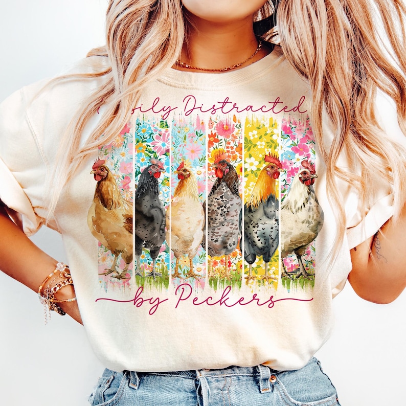 May include: Cream-colored t-shirt featuring a colorful floral design with six chickens. The text "Totally Distracted by Peckers" is printed above and below the chickens. The shirt is made of a soft material.