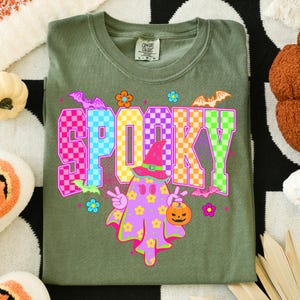 May include: Olive green t-shirt with the word "SPOOKY" in a checkered pattern. The shirt features a cartoon ghost wearing a witch hat and holding a jack-o'-lantern. Bats and flowers are also part of the design, perfect for Halloween.
