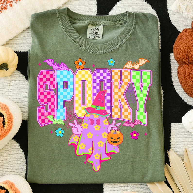 May include: Olive green t-shirt with the word "SPOOKY" in a checkered pattern. The shirt features a cartoon ghost wearing a witch hat and holding a jack-o'-lantern. Bats and flowers are also part of the design, perfect for Halloween.