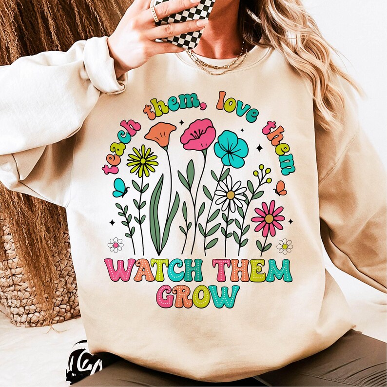 Puede incluir: Una sudadera beige con un dise&ntilde;o gr&aacute;fico colorido que presenta flores y el texto "teach them, love them, watch them grow".