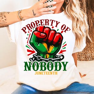 Może przedstawiać: Biała koszulka z motywem zaciśniętej pięści w kolorze czerwonym, zielonym i ż&oacute;łtym, z napisami "PROPERTY OF NOBODY" i "JUNETEENTH" w kolorze zielonym i czerwonym. Pięść jest zakuta w łańcuch. Koszulka to element odzieży codziennej.