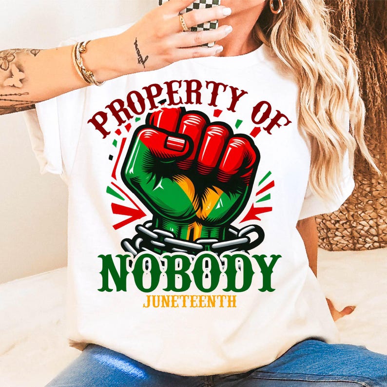 Może przedstawiać: Biała koszulka z motywem zaciśniętej pięści w kolorze czerwonym, zielonym i ż&oacute;łtym, z napisami "PROPERTY OF NOBODY" i "JUNETEENTH" w kolorze zielonym i czerwonym. Pięść jest zakuta w łańcuch. Koszulka to element odzieży codziennej.