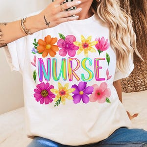 以下が含まれることがあります： カラフルな花の模様と、虹色のグラデーションフォントで「NURSE」の文字が入った白いTシャツ。花はオレンジ、ピンク、黄色、紫の色合いです。カジュアルな普段着です。