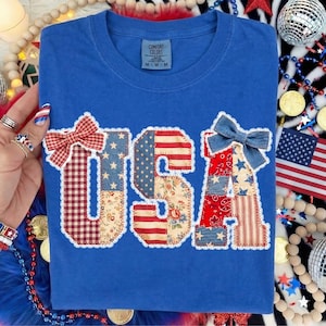 May include: A blue t-shirt with the word "USA" appliqued on the front. The letters are made of patchwork fabric in red, white, and blue patterns, with a bow on the "U" and "A". The shirt has a crew neck and a tag that says "Comfort Colors".