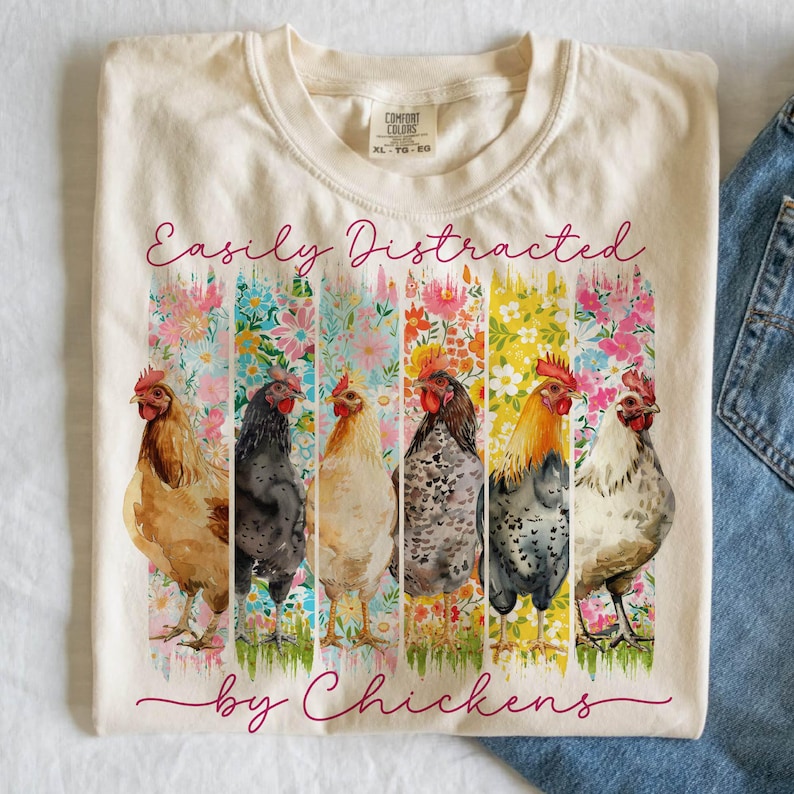 以下が含まれることがあります： クリーム色のTシャツ。筆記体で「Easily Distracted by Chickens」と書かれています。シャツには、花の背景の前に6羽の鶏の水彩デザインが施されています。シャツはComfort Colors XL-TG-EGです。
