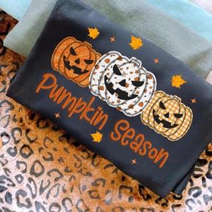 May include: A black sweatshirt featuring three jack-o'-lanterns in orange, white, and plaid patterns. The phrase "Pumpkin Season" is written in orange script. Autumn leaves and stars are scattered around the design.