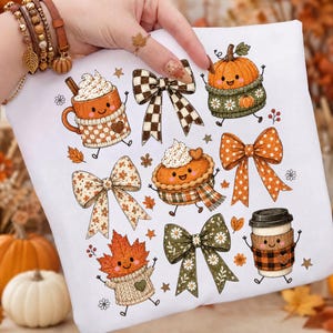 May include: A white t-shirt featuring a colorful autumn-themed graphic print. The design includes smiling anthropomorphic characters such as a pumpkin in a sweater, a coffee cup, a pie, and a maple leaf, interspersed with various patterned bows.