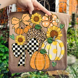 以下が含まれることがあります： 秋をテーマにしたデザインの茶色の装飾品。デザインには、オレンジ、黄色、茶色のカボチャ、ヒマワリ、リボンが含まれています。カボチャの1つはヒョウ柄です。緑のハートのアクセントがデザインを完成させています。