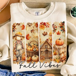 Puede incluir: Sudadera de color crema con un gráfico temático de otoño. El diseño presenta un espantapájaros, calabazas, una cesta de calabazas, una casa y hojas de otoño. Las palabras "Fall Vibes" están escritas en una fuente cursiva.