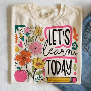 May include: Cream-colored t-shirt with the words "LET'S learn TODAY" in black script, framed by a pink rectangle. The shirt features colorful floral and apple graphics, and a yellow pencil with a pink eraser. The shirt is paired with blue jeans.