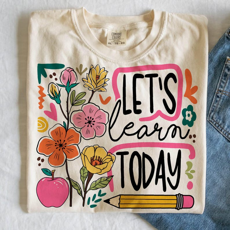 May include: Cream-colored t-shirt with the words "LET'S learn TODAY" in black script, framed by a pink rectangle. The shirt features colorful floral and apple graphics, and a yellow pencil with a pink eraser. The shirt is paired with blue jeans.