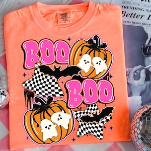 Puede incluir: Camiseta naranja Comfort Colors con un dise&ntilde;o de Halloween. El dise&ntilde;o incluye la palabra "BOO" en letras rosas, calabazas, fantasmas, murci&eacute;lagos y estrellas a cuadros. La camiseta es talla L-G.