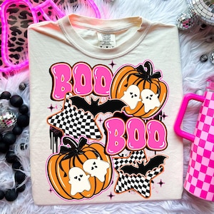 Puede incluir: Camiseta color crema con un dise&ntilde;o tem&aacute;tico de Halloween. El dise&ntilde;o incluye la palabra "BOO" en rosa, calabazas naranjas, fantasmas blancos, murci&eacute;lagos negros y estrellas a cuadros. La camiseta es de la marca Comfort Colors.