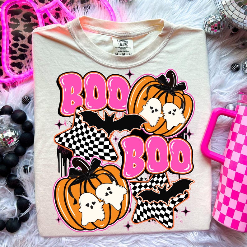Puede incluir: Camiseta color crema con un dise&ntilde;o tem&aacute;tico de Halloween. El dise&ntilde;o incluye la palabra "BOO" en rosa, calabazas naranjas, fantasmas blancos, murci&eacute;lagos negros y estrellas a cuadros. La camiseta es de la marca Comfort Colors.