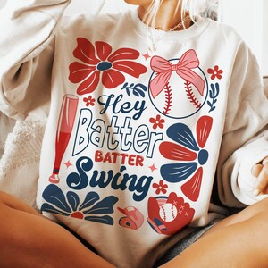 May include: A white sweatshirt with a baseball-themed design. It features a red, white, and blue color scheme with flowers, a baseball, a bat, and the text "Hey Batter Batter Swing".