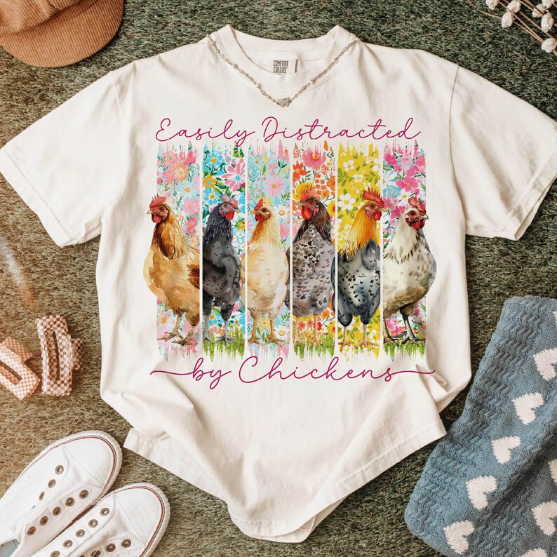 以下が含まれることがあります： 「Easily Distracted by Chickens」の文字が入った白いTシャツ。6羽の鶏が花柄の背景の前に描かれた、カラフルなグラフィックが特徴です。