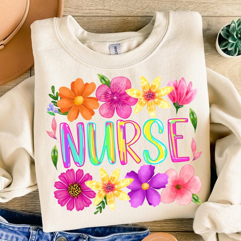 以下が含まれることがあります： クリーム色のスウェットシャツ。カラフルなバブル文字で「NURSE」の文字が描かれ、オレンジ、ピンク、黄色、紫の色合いの水彩画の花々が周りを囲んでいます。緑の葉とアクセントが施され、陽気で花柄のテーマを作り出しています。