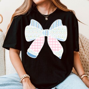 May include: A black t-shirt featuring a large, colourful bow design. The bow is made from various patterned fabrics, including pink and blue gingham, floral print, and stripes.
