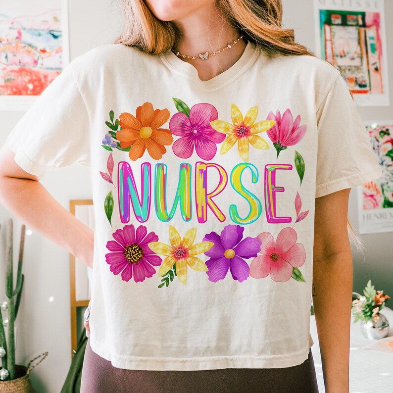 以下が含まれることがあります： クリーム色のTシャツ。レインボーカラーの「NURSE」の文字がプリントされ、オレンジ、ピンク、黄色、紫色の水彩画の花で囲まれています。 ゆったりとしたフィット感で、カジュアルなスタイルです。