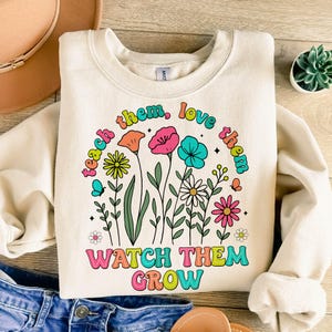 Puede incluir: Una sudadera de color crema con un dise&ntilde;o floral colorido y el texto "Teach them, love them, watch them grow".