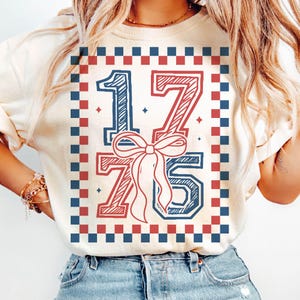 May include: Cream-colored sweatshirt with a patriotic design. The graphic features the numbers "1775" in red and blue, with a bow in the center. The design is framed by a red and blue checkered border. The shirt is paired with blue jeans.