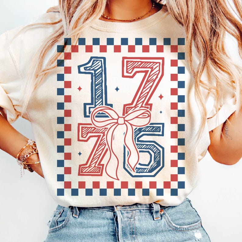 May include: Cream-colored sweatshirt with a patriotic design. The graphic features the numbers "1775" in red and blue, with a bow in the center. The design is framed by a red and blue checkered border. The shirt is paired with blue jeans.
