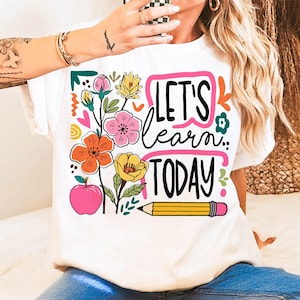 May include: White t-shirt with a colorful floral design and the text "LET'S learn TODAY". The design includes orange, pink, and yellow flowers, a pink apple, and a yellow pencil. A fun and educational theme.