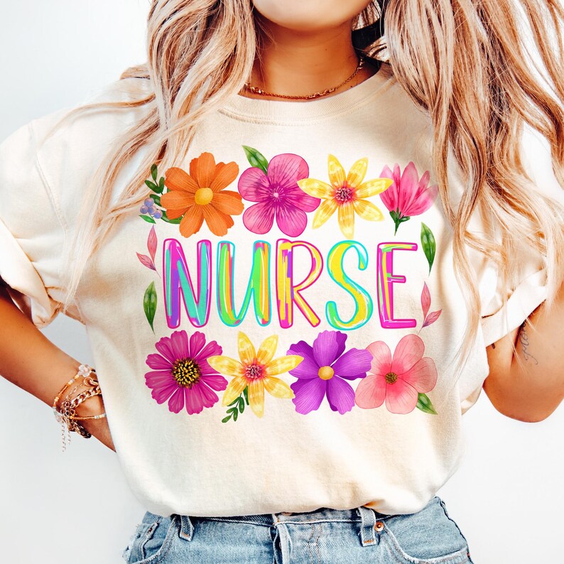以下が含まれることがあります： クリーム色のTシャツ。カラフルな虹色の文字で「NURSE」と書かれており、オレンジ、ピンク、黄色、紫の色合いの鮮やかな花のイラストで囲まれています。このシャツはクルーネックで半袖です。