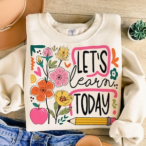 May include: Cream-colored sweatshirt with a colorful floral design and the text "LET'S learn TODAY". The graphic includes a pink apple, a yellow pencil, and various flowers in pink, orange, and yellow.