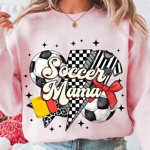K&ouml;nnte beinhalten: Rosafarbener Pullover mit einem Grafikdesign aus einem Fu&szlig;ball, einem Schiedsrichtertrikot und einem karierten Muster. Der Text "Soccer Mama" ist in einer Retro-Schrift geschrieben.