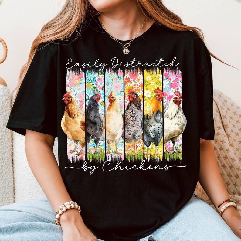 以下が含まれることがあります： 「Easily Distracted by Chickens」の文字が入った黒いTシャツ。カラフルな花柄のデザインと、6羽の異なる鶏のイラストが特徴です。鶏は茶色、黒、白など、さまざまな色をしています。