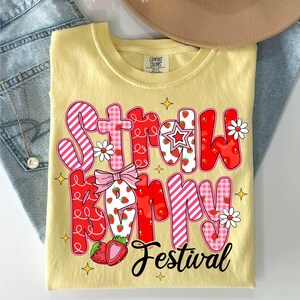 May include: A yellow t-shirt with a red and pink graphic that says "Strawberry Festival". The graphic is made up of letters with a strawberry and gingham pattern.