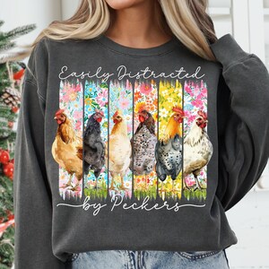 May include: Dark gray sweatshirt with the text "Easily Distracted by Peckers." The graphic features six colorful chickens against a floral background. The chickens are in various colors, including brown, black, and white.