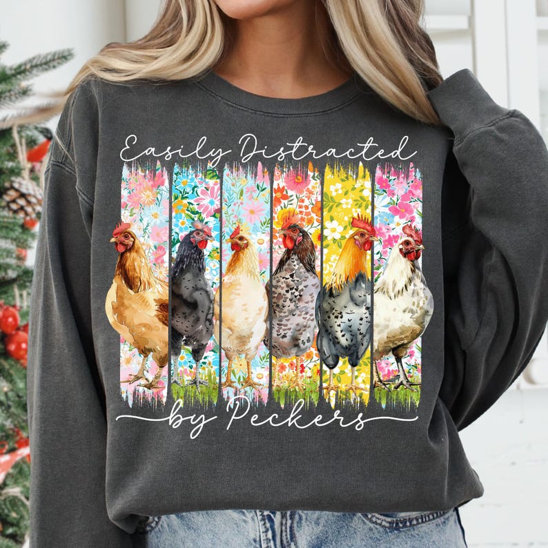 May include: Dark gray sweatshirt with the text "Easily Distracted by Peckers." The graphic features six colorful chickens against a floral background. The chickens are in various colors, including brown, black, and white.