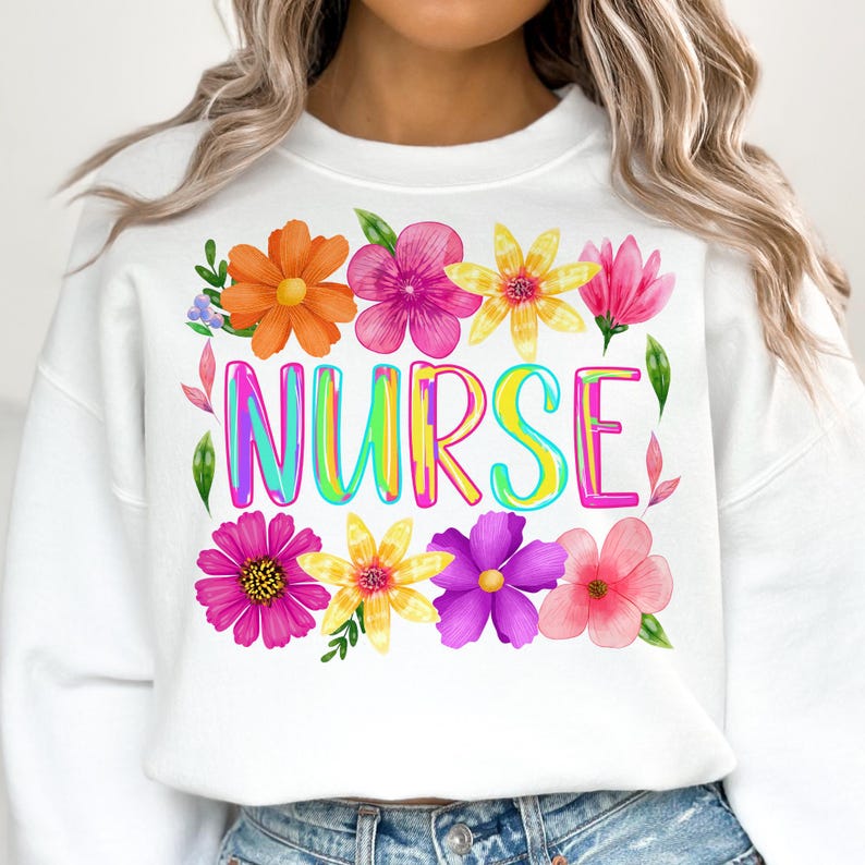 以下が含まれることがあります： カラフルな文字で「NURSE」と書かれた白いスウェットシャツ。オレンジ、ピンク、黄色、紫の色合いの水彩画の花で囲まれています。デザインには緑の葉とアクセントが含まれており、陽気で花柄のテーマを作り出しています。