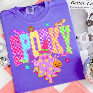 May include: A purple t-shirt with the word "SPOOKY" in colorful checkered letters. A cartoon ghost with a witch hat and a pumpkin bucket is in the center. Bats and flowers decorate the shirt, perfect for Halloween.