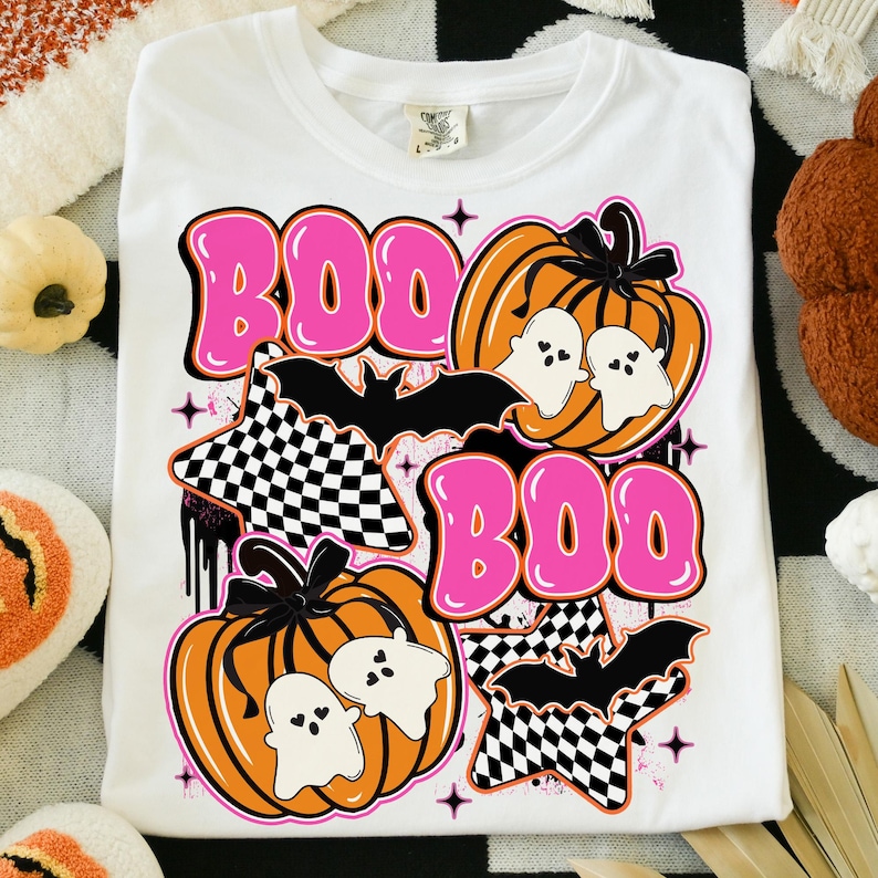 Puede incluir: Camiseta blanca con un dise&ntilde;o tem&aacute;tico de Halloween. El dise&ntilde;o incluye la palabra "BOO" en letras rosas, calabazas naranjas, fantasmas blancos, murci&eacute;lagos negros y patrones de cuadros. La camiseta est&aacute; doblada.