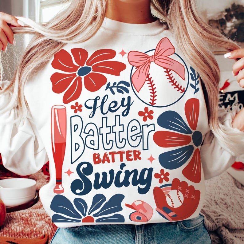 May include: White sweatshirt with a baseball theme. It features a red, white, and blue floral design, a baseball, a bat, and the text "Hey Batter Batter Swing".