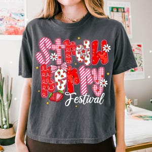 May include: A gray t-shirt with a graphic design of the words "Strawberry Festival" in pink, red, and white with a strawberry pattern. The words are in a patchwork style with a bow and stars.
