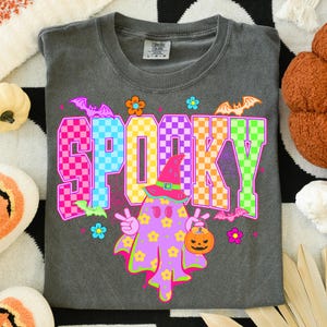 May include: A gray Comfort Colors t-shirt featuring the word "SPOOKY" in a colorful checkered design. A cartoon ghost with a witch hat and a pumpkin bucket is in the center. Bats and flowers surround the text, creating a festive Halloween theme.